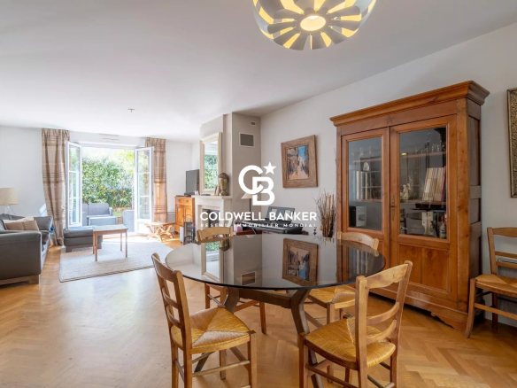 Bright open-plan living and dining room with a round glass table and wooden chairs, beige sofa, and a large wooden cabinet, leading to a garden through open French doors, with a modern ceiling light and parquet floor. The Coldwell Banker logo is centered on the image.