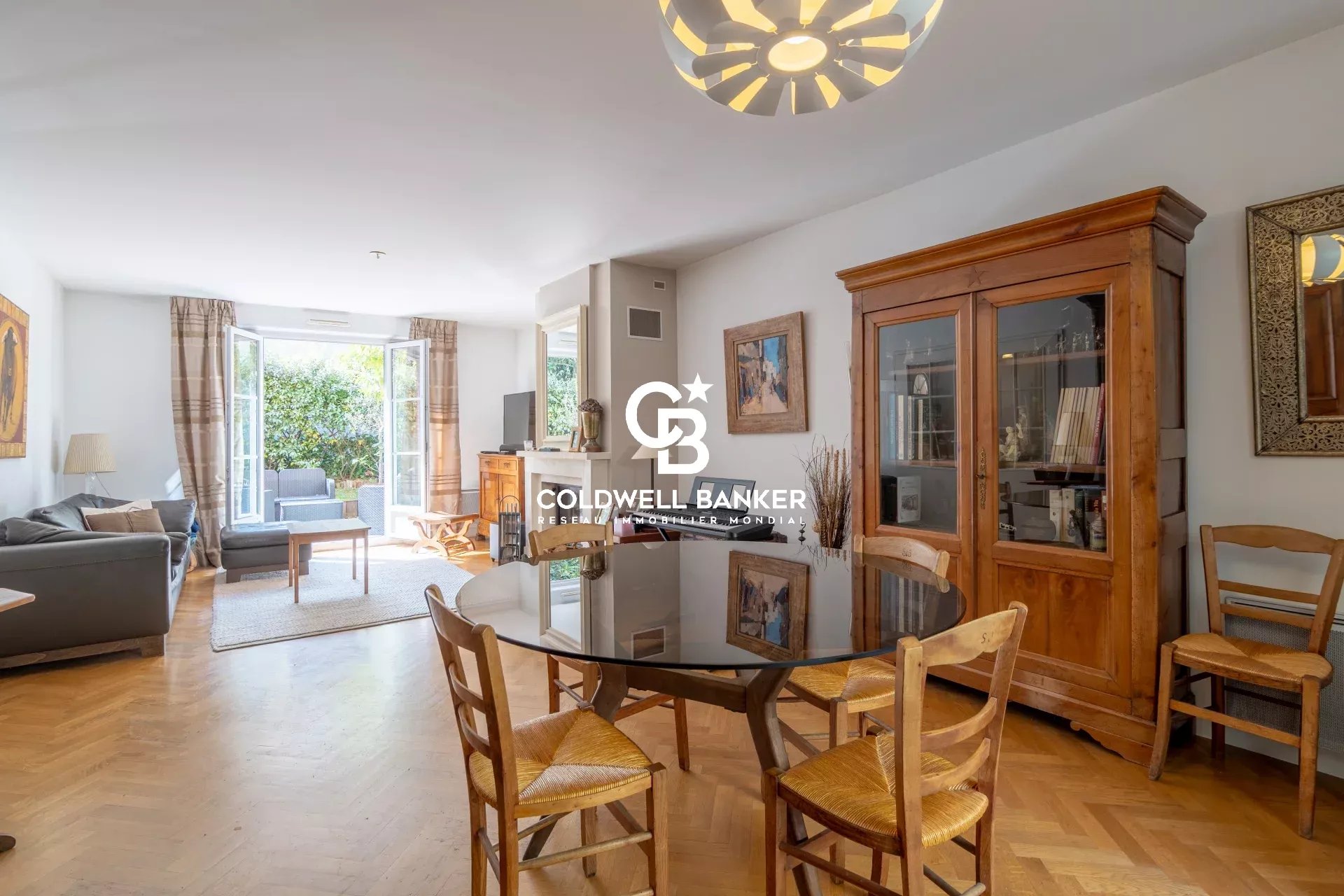 Bright open-plan living and dining room with a round glass table and wooden chairs, beige sofa, and a large wooden cabinet, leading to a garden through open French doors, with a modern ceiling light and parquet floor. The Coldwell Banker logo is centered on the image.