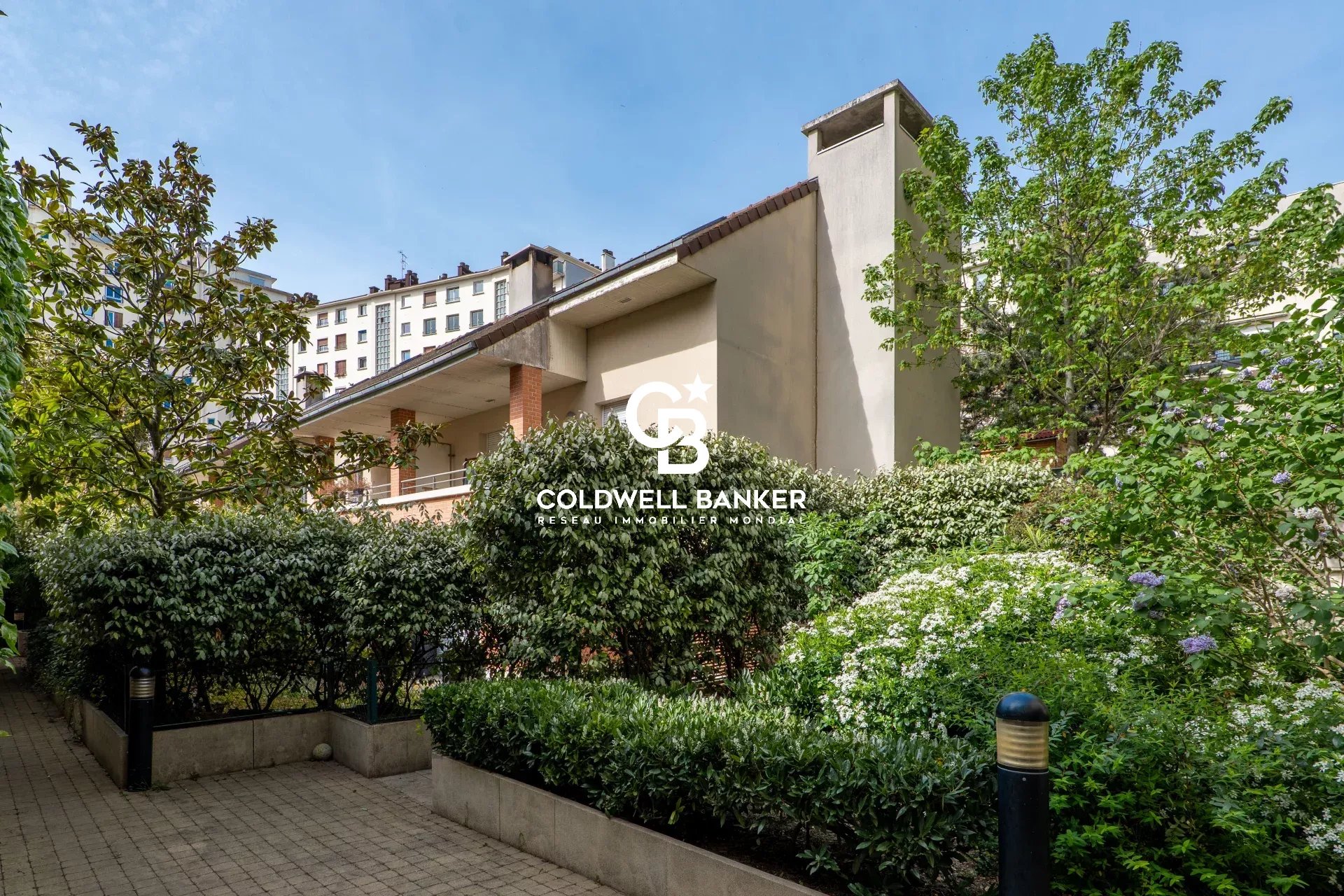 Residential building with a manicured garden, hedges, and a paved walkway under a blue sky; Coldwell Banker logo overlayed in the center.