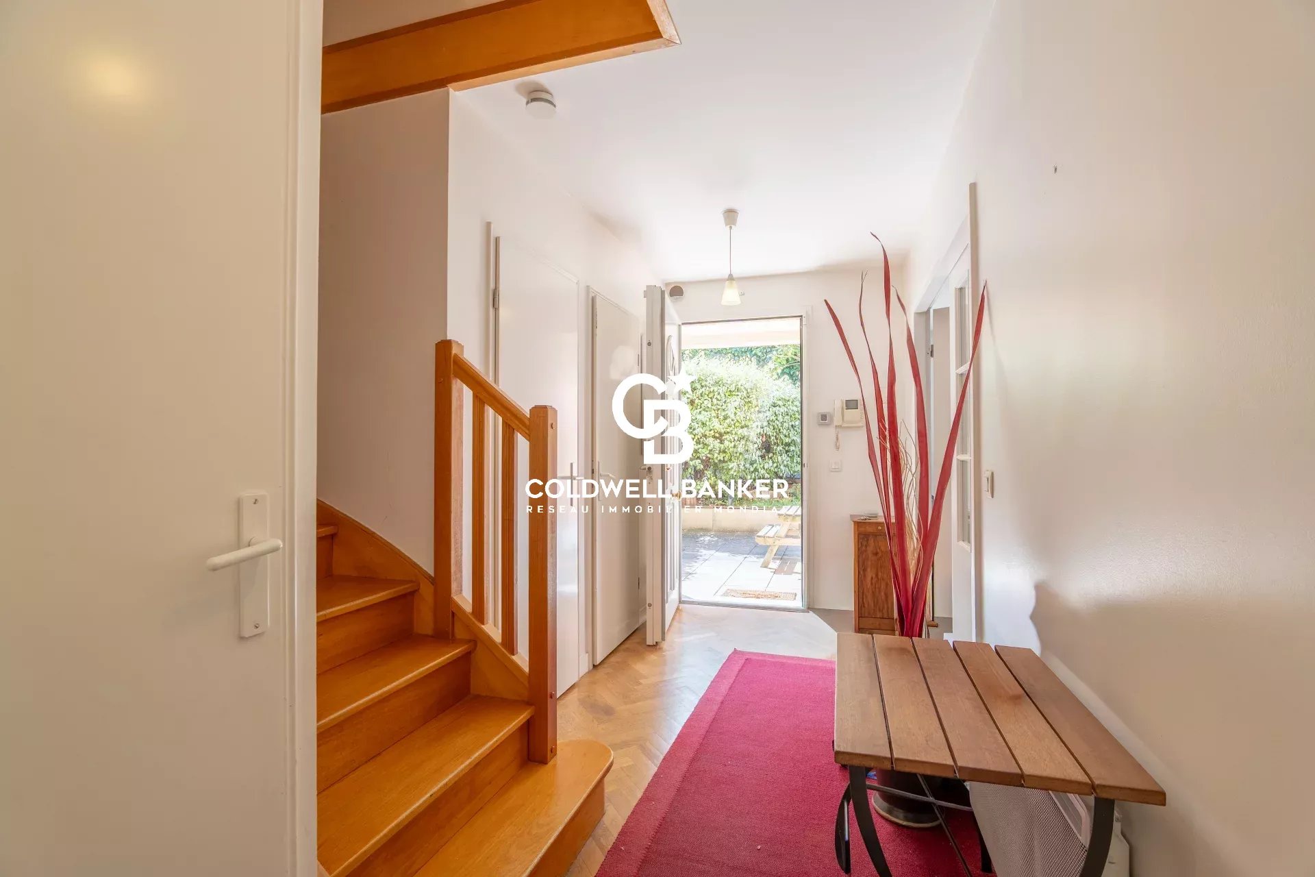 Entry hallway with a wooden staircase on the left, a red runner rug, and a glass door opening to a sunny garden.