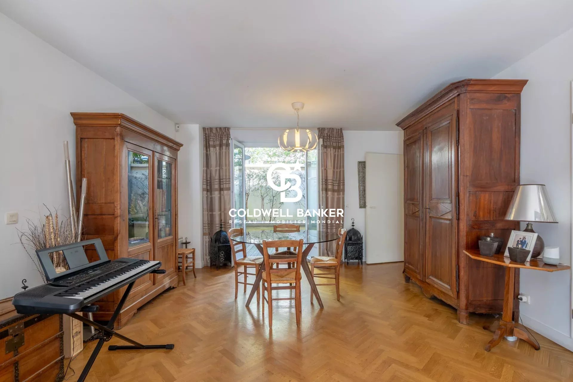 Living room with parquet floor, two large wooden wardrobes, a glass-front cabinet, a round dining table with four chairs, and a keyboard on a stand by the left wall.