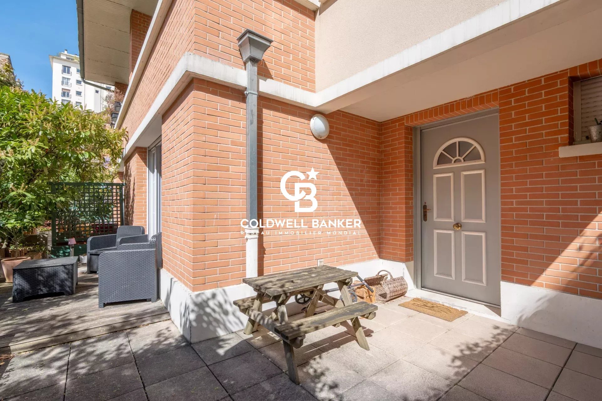 Brick exterior with a small patio featuring wicker seating and a wooden picnic table, adjacent to a gray front door with a semicircular window panel.