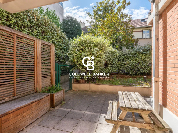 Patio area with a wooden bench, a weathered picnic table, and green hedges along a brick wall, featuring a Coldwell Banker logo in the center.