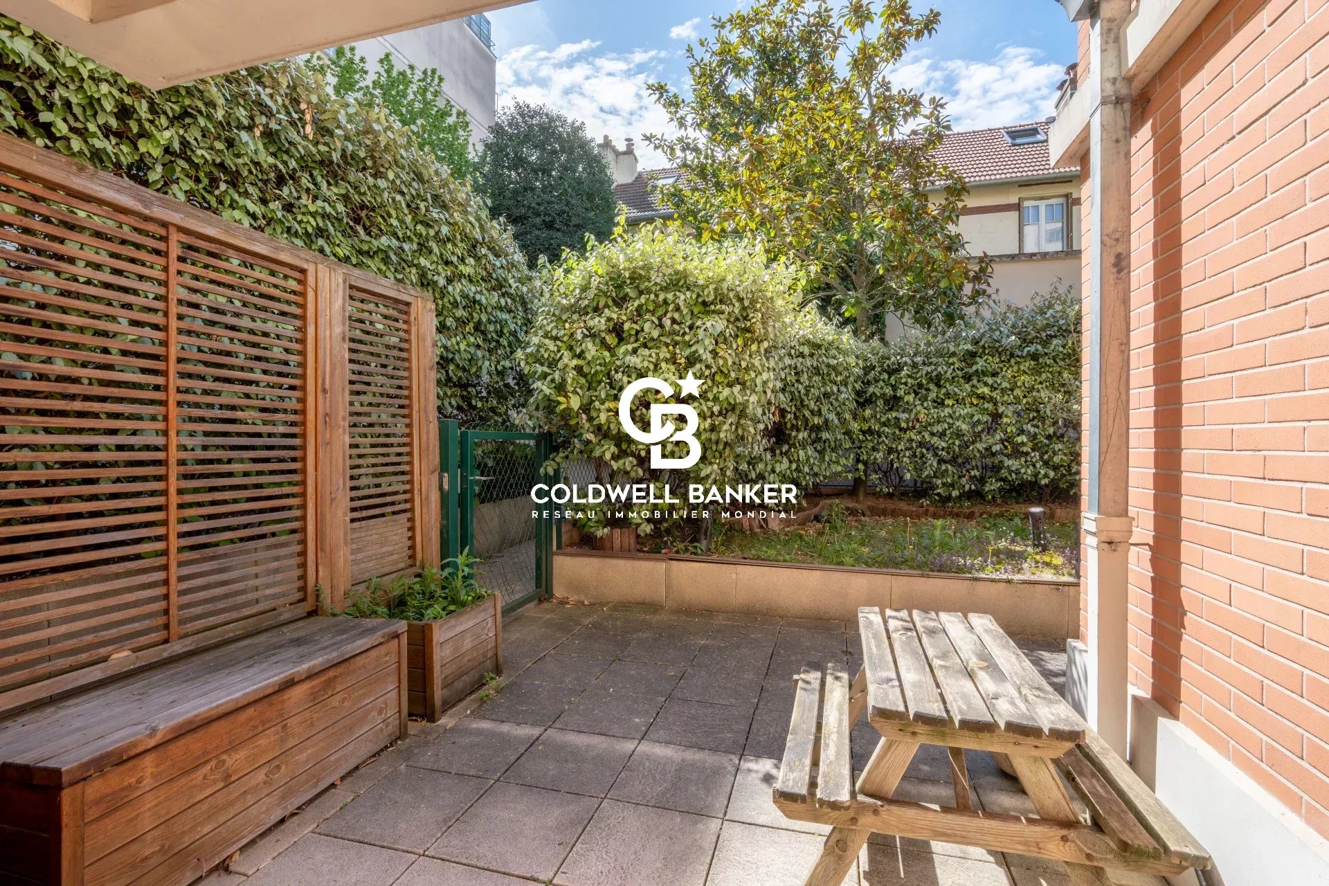 Patio area with a wooden bench, a weathered picnic table, and green hedges along a brick wall, featuring a Coldwell Banker logo in the center.