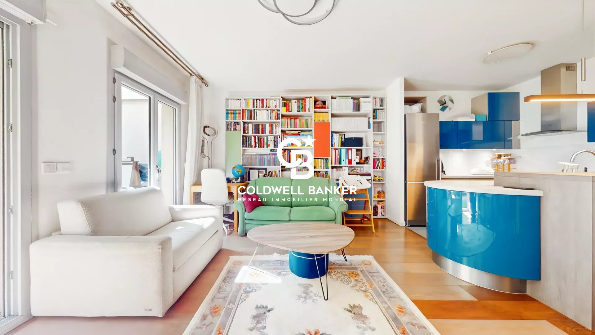 Open-plan living area with a white sofa, green chair, and a tall bookcase wall, leading to a blue curved kitchen island and fridge; watermark in center.
