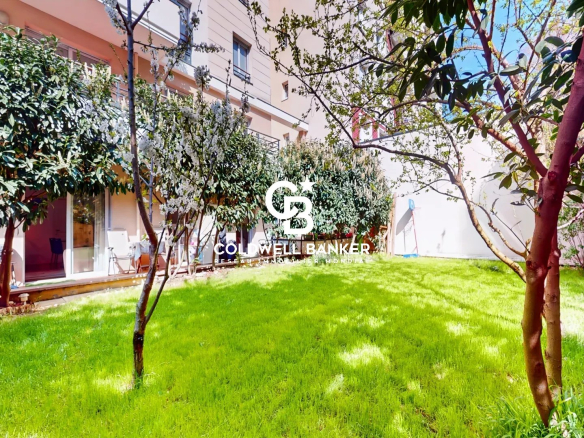 Sunny residential courtyard with a well-kept green lawn, trees, and a glass-walled patio area along the building; Coldwell Banker logo overlay.