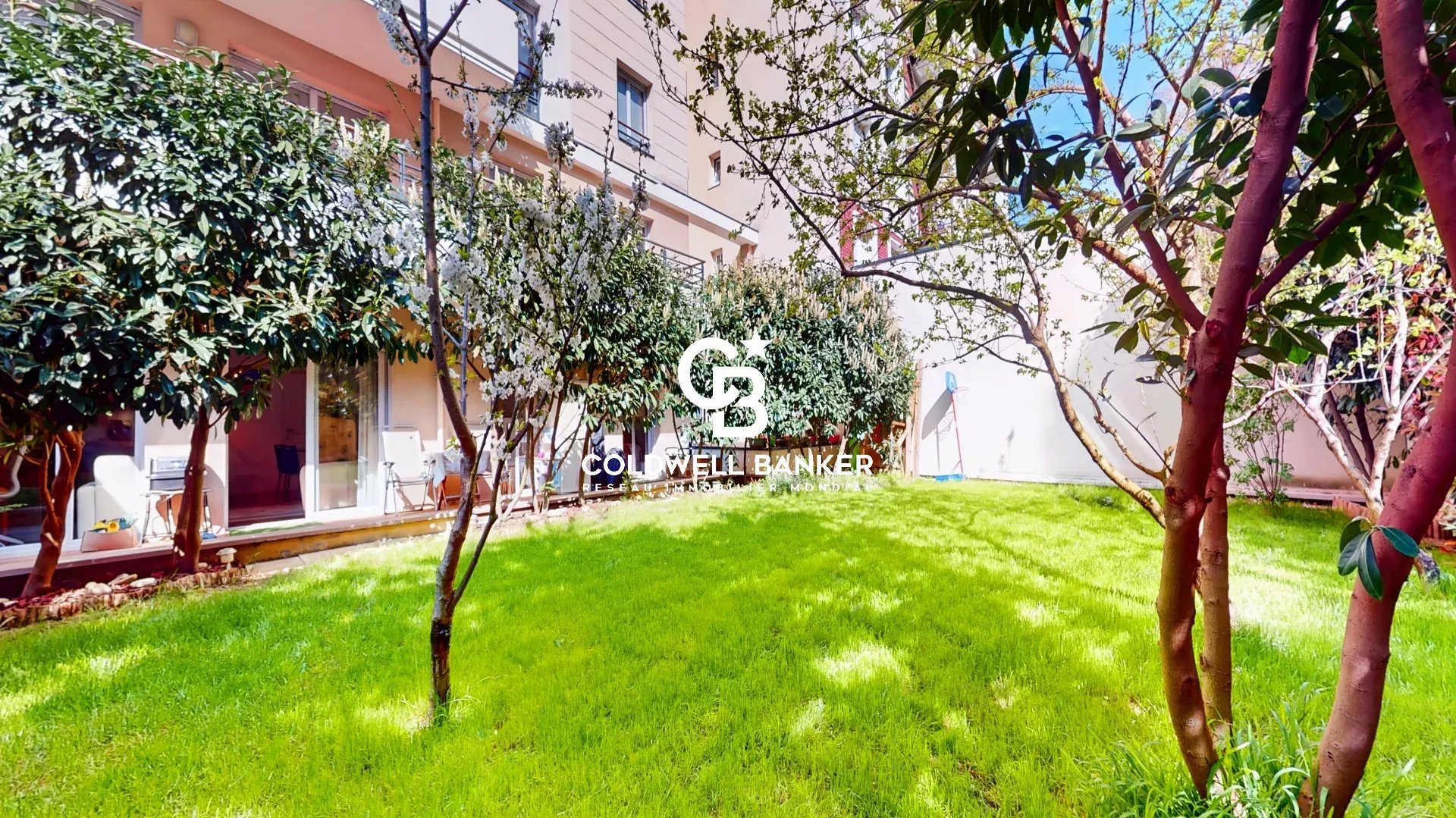 Sunny residential courtyard with a well-kept green lawn, trees, and a glass-walled patio area along the building; Coldwell Banker logo overlay.