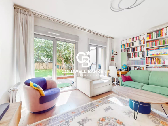 Bright living room with a green sofa, two white armchairs, and a wall of colorful bookshelves facing large windows showing a green garden outside.
