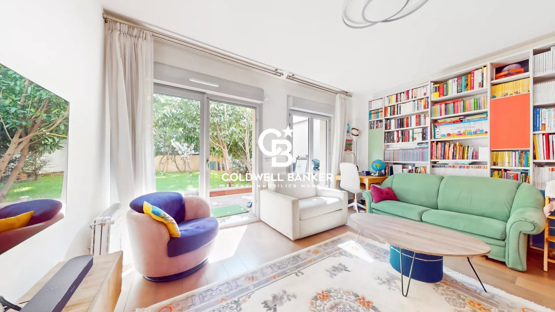 Bright living room with a green sofa, two white armchairs, and a wall of colorful bookshelves facing large windows showing a green garden outside.
