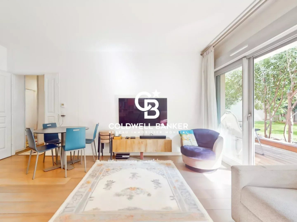 Bright open-plan living room with a round dining table and blue chairs, TV mounted on the wall, and sliding glass doors to a green garden outside, featuring a light rug and neutral sofa.