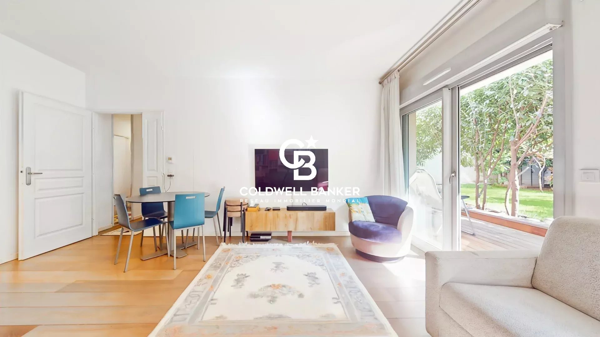 Bright open-plan living room with a round dining table and blue chairs, TV mounted on the wall, and sliding glass doors to a green garden outside, featuring a light rug and neutral sofa.