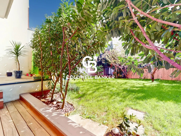 Sunlit backyard with a wooden deck, steps, and potted plants along a white wall, leading to a lush green lawn. Watermark: Coldwell Banker.