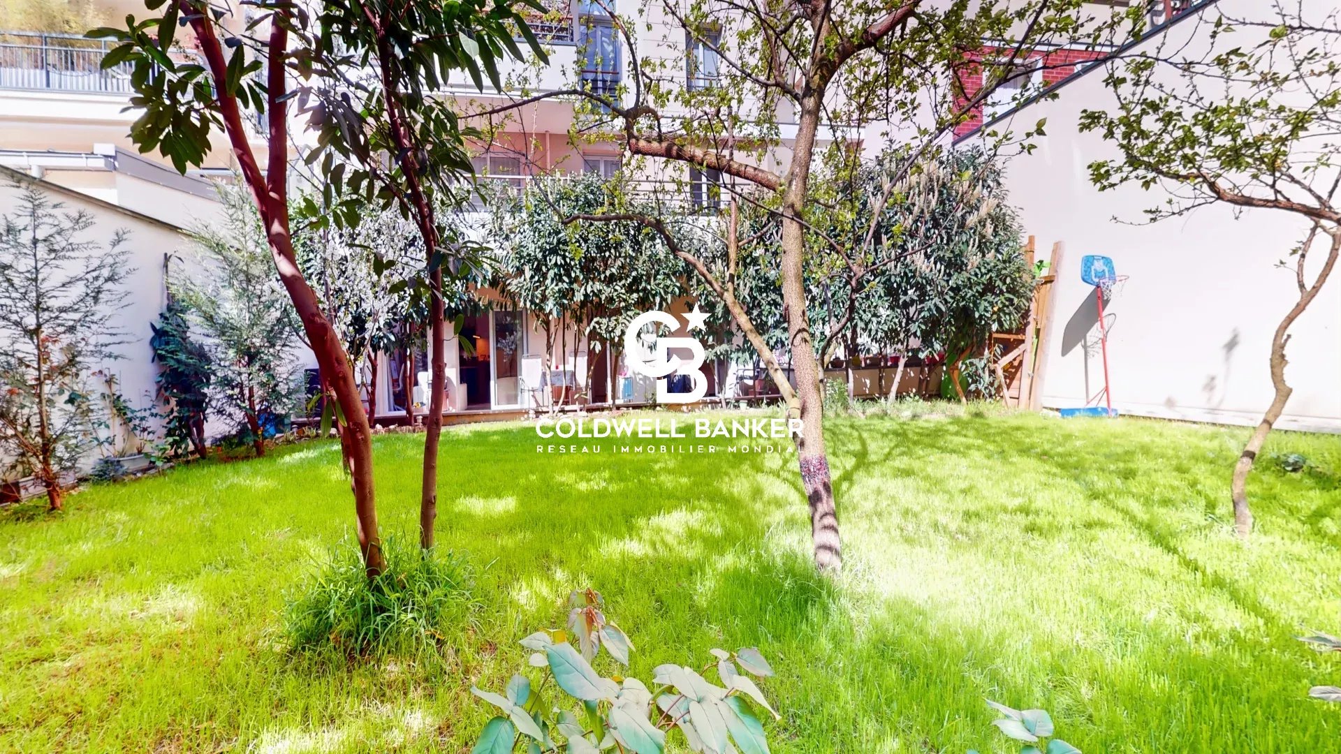 Green grassy courtyard with several trees and shrubs, bordered by white walls and apartment balconies, a small basketball hoop on the right.