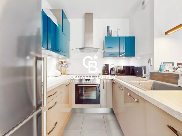 Modern L-shaped kitchen with blue upper cabinets, stainless steel appliances, and light wood lower cabinets under a white countertop.