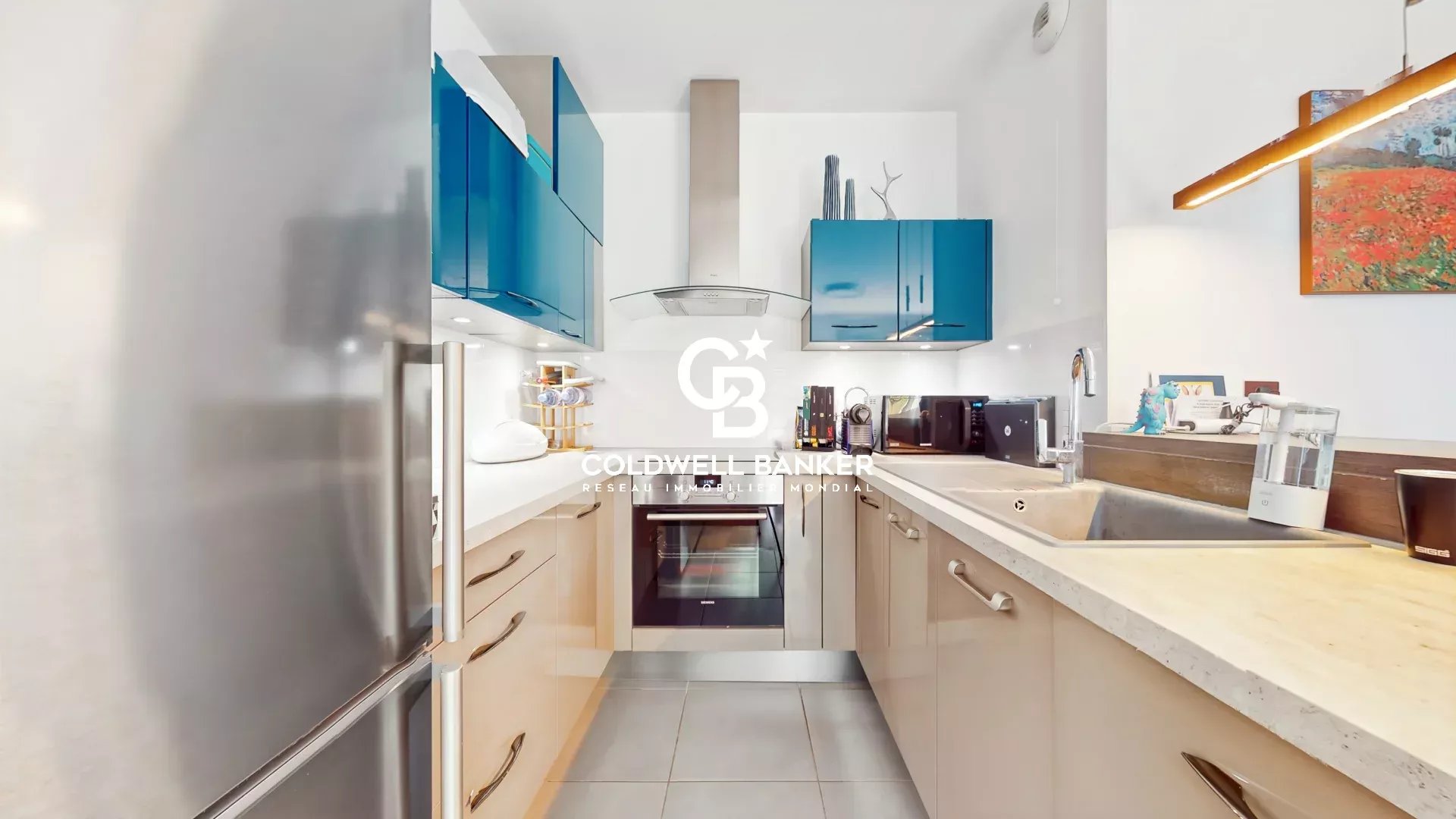 Modern L-shaped kitchen with blue upper cabinets, stainless steel appliances, and light wood lower cabinets under a white countertop.