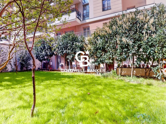 Courtyard garden of a modern apartment building with lush green lawn, trees, and hedges; watermark reads 'Goldwell Banker' in the center.