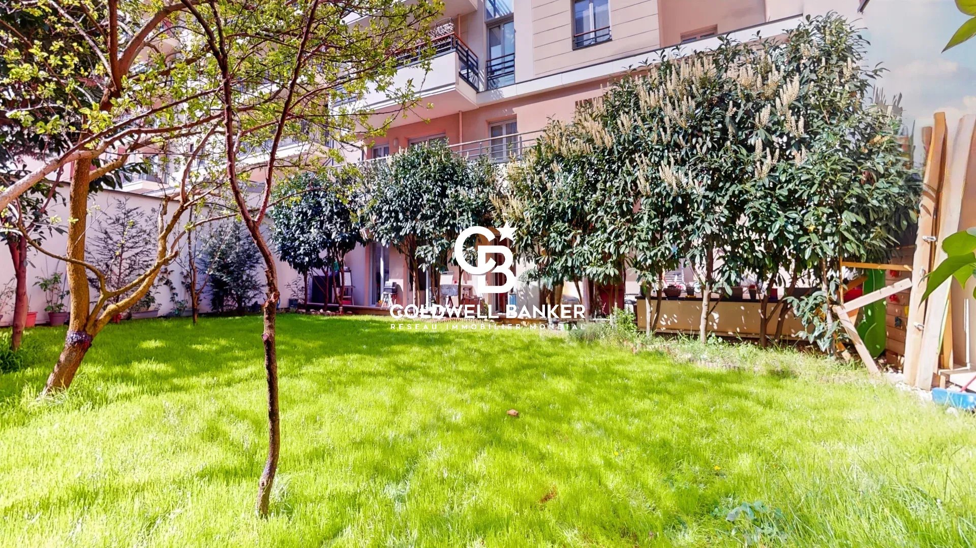 Courtyard garden of a modern apartment building with lush green lawn, trees, and hedges; watermark reads 'Goldwell Banker' in the center.