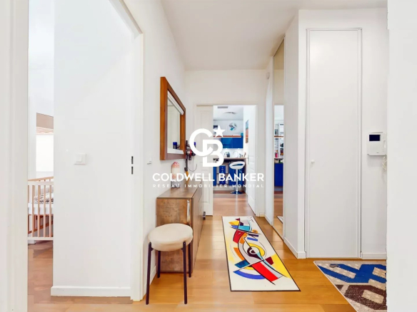 Bright hallway in a modern home with white walls and warm wooden flooring, a small bench and round stool on the left, and a colorful abstract rug leading toward a blue-accented kitchen beyond. The image has a Coldwell Banker watermark in the center.