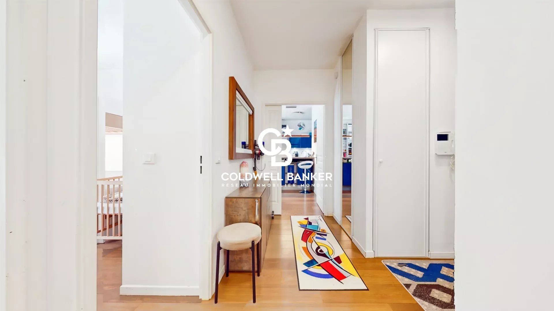 Bright hallway in a modern home with white walls and warm wooden flooring, a small bench and round stool on the left, and a colorful abstract rug leading toward a blue-accented kitchen beyond. The image has a Coldwell Banker watermark in the center.