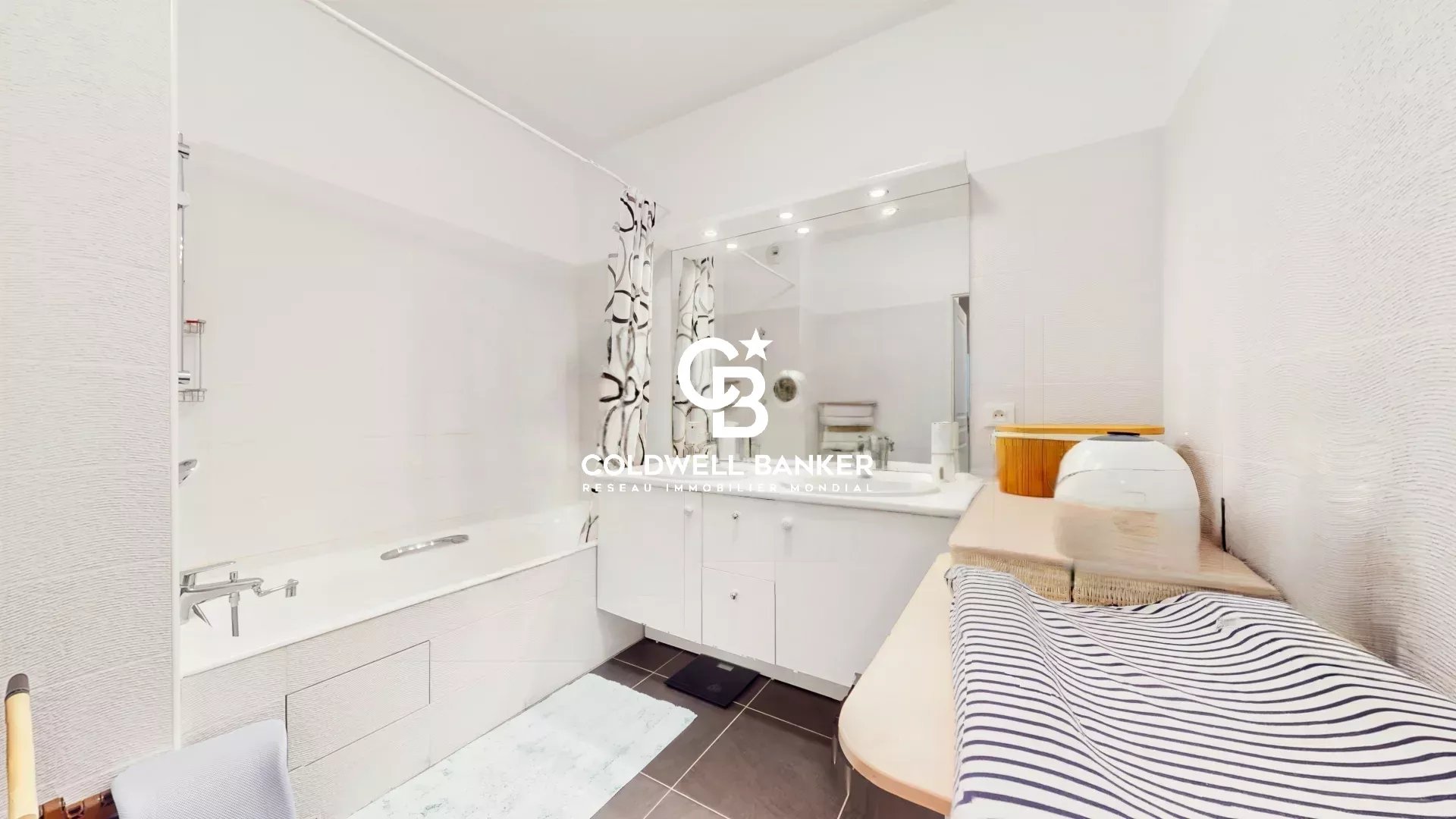 Modern white bathroom with bathtub, vanity and large mirror; Coldwell Banker logo centered over the scene.