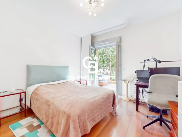 Bright bedroom with a queen bed, pale teal headboard, and peach bedspread; a small red side table and shelf to the left, wooden floor, and a balcony door to the outside on the right; a workspace with a monitor and desk chair on the far right.