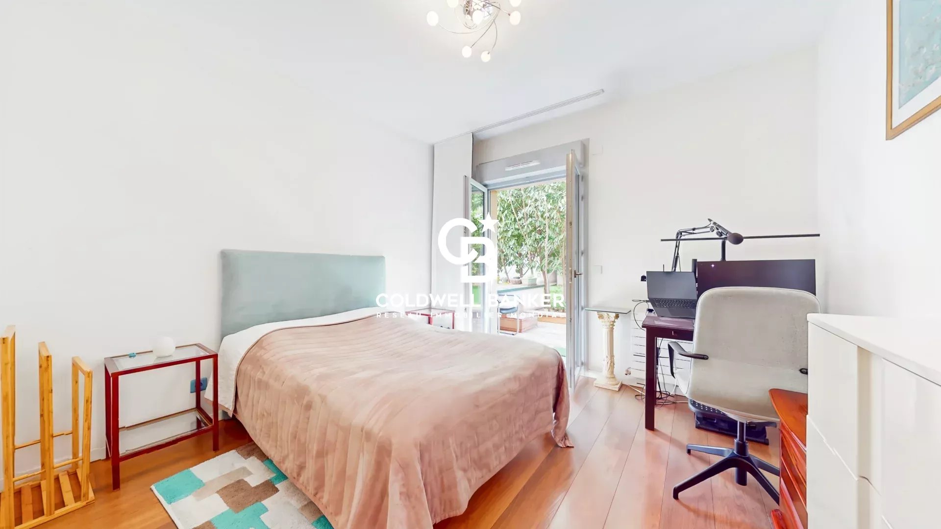 Bright bedroom with a queen bed, pale teal headboard, and peach bedspread; a small red side table and shelf to the left, wooden floor, and a balcony door to the outside on the right; a workspace with a monitor and desk chair on the far right.
