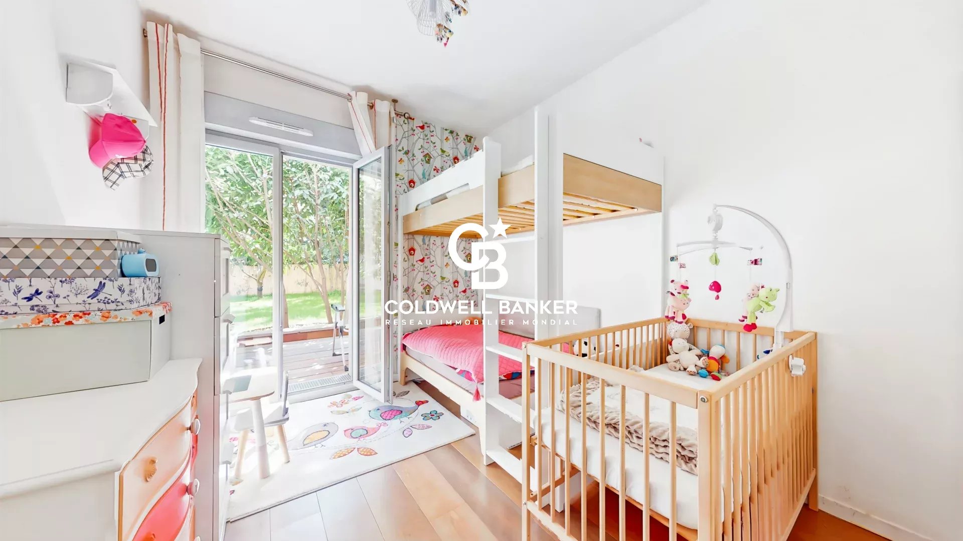 Bright children's bedroom with white walls, a wooden bunk bed and a wooden crib, and a white dresser with colorful drawers near sliding glass doors to a green garden exterior
