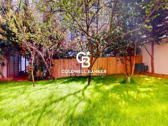 Sunlit backyard with a lush green lawn, trees, and a wooden fence; Coldwell Banker branding centered in the image.