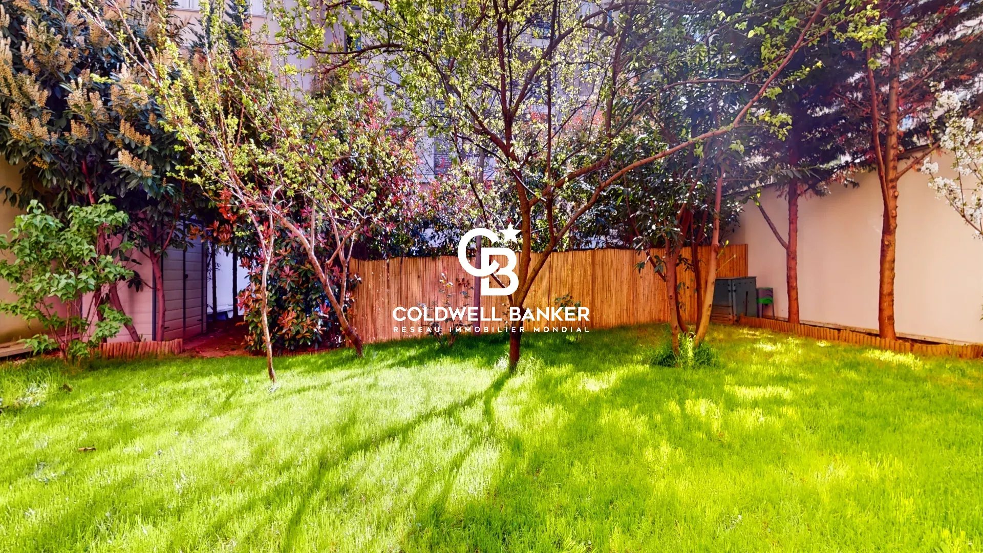 Sunlit backyard with a lush green lawn, trees, and a wooden fence; Coldwell Banker branding centered in the image.