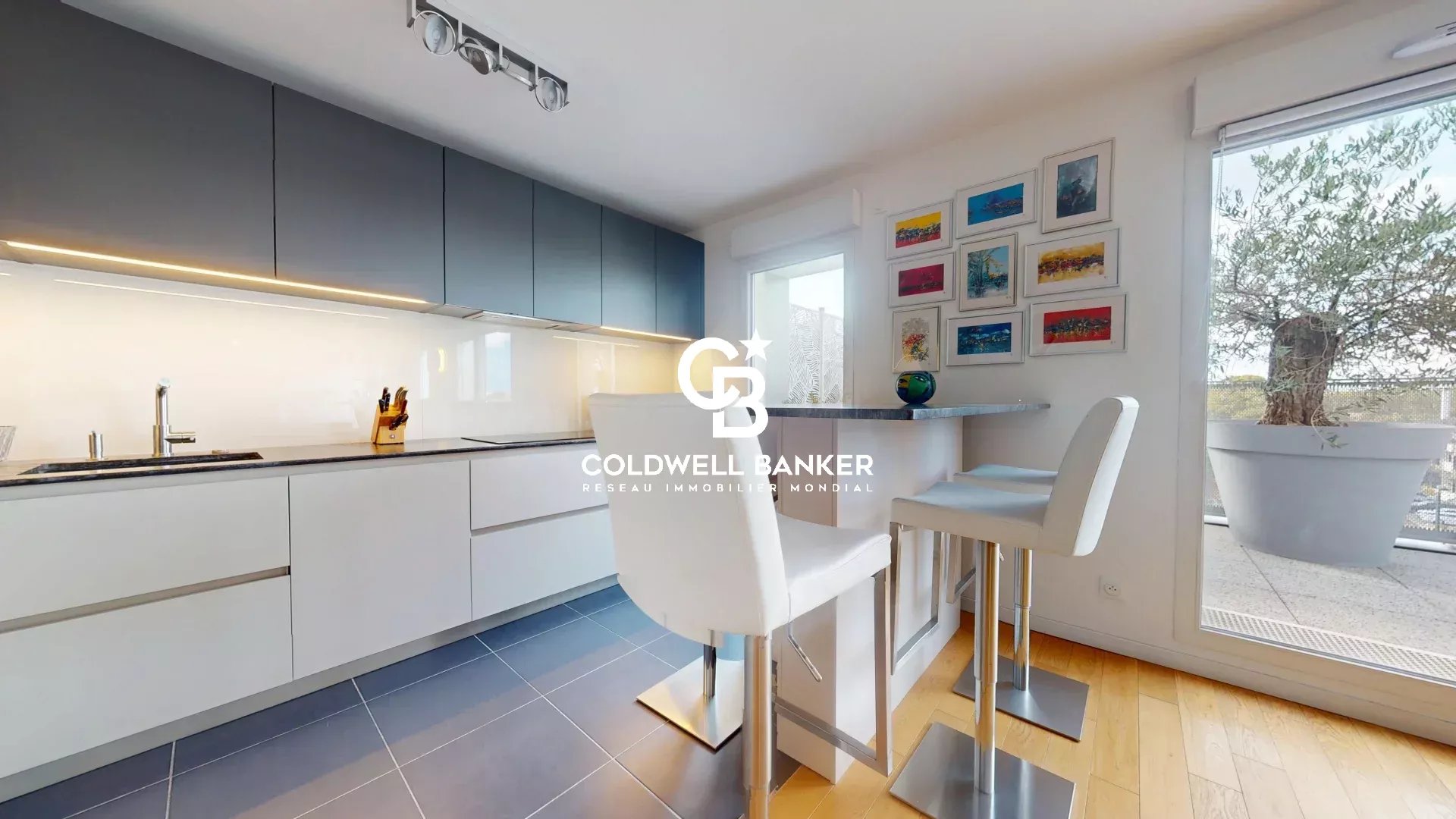 Cuisine moderne d'un appartement avec bar et chaises hautes, fenêtres donnant sur une terrasse.