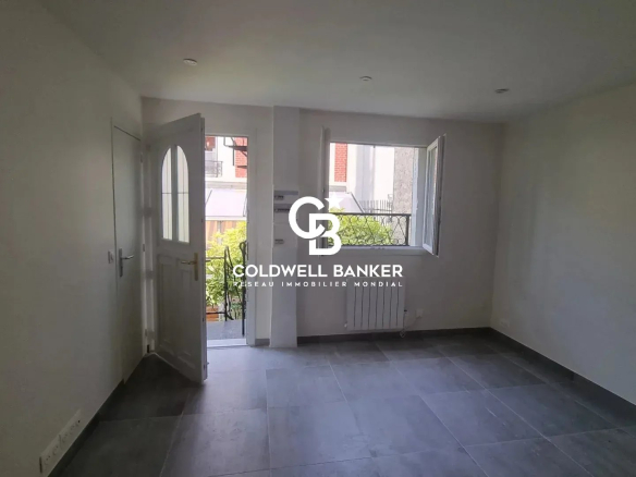 Intérieur d'un studio rénové avec fenêtre et porte d'entrée, sol en carrelage gris.