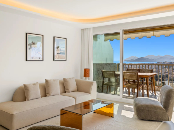 Bright living room with beige sectional sofa, matching pillows, and a glass amber coffee table facing a sliding glass door to a balcony. Balcony has a wooden table and chairs with a ocean and mountain view.