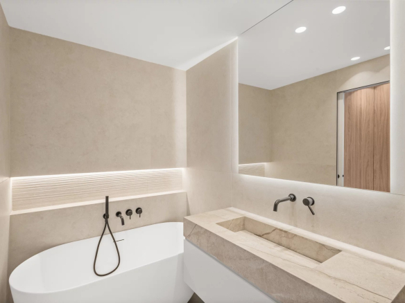 Modern beige-tiled bathroom with a freestanding white bathtub, wall-mounted black fixtures, and a large backlit mirror.