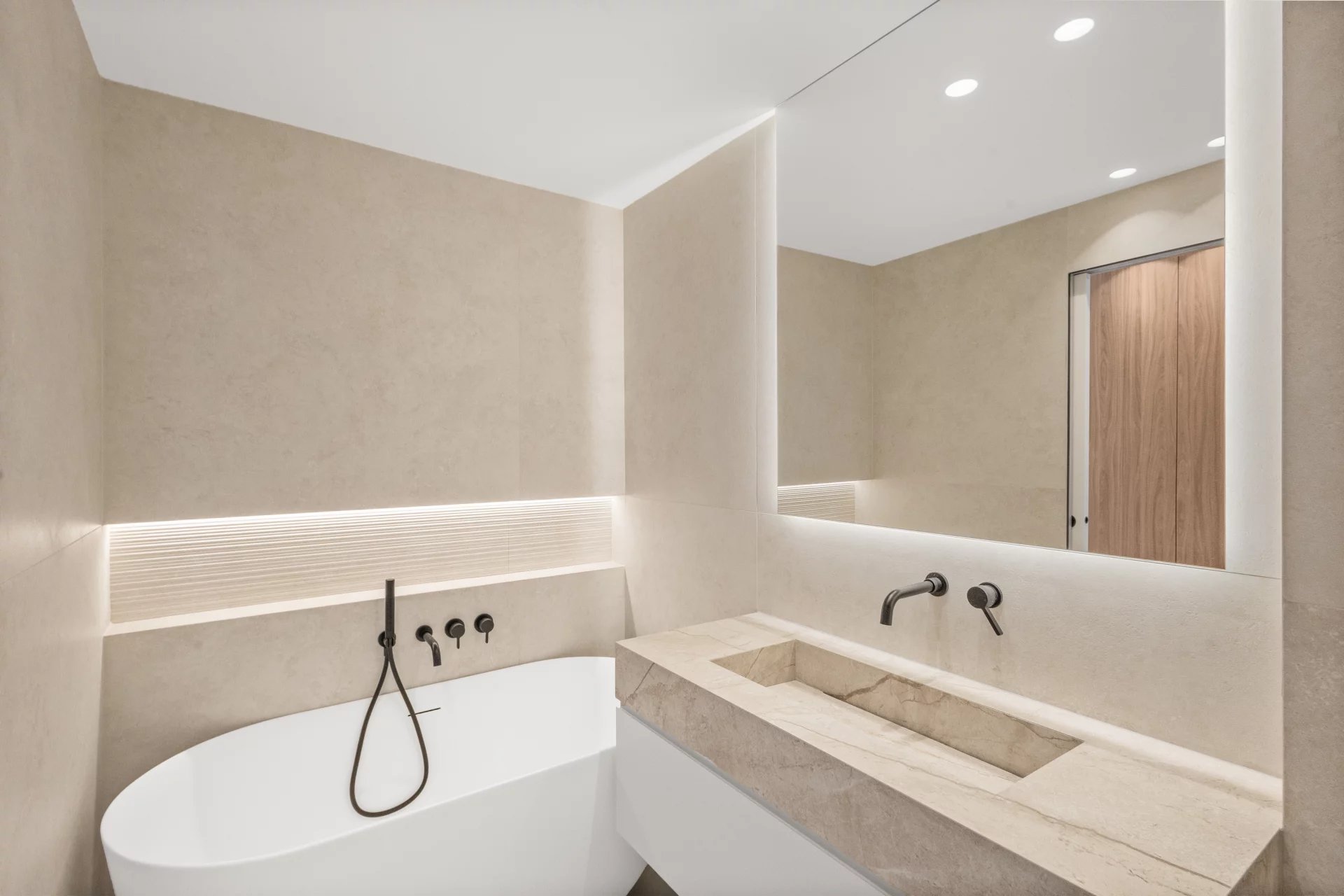 Modern beige-tiled bathroom with a freestanding white bathtub, wall-mounted black fixtures, and a large backlit mirror.
