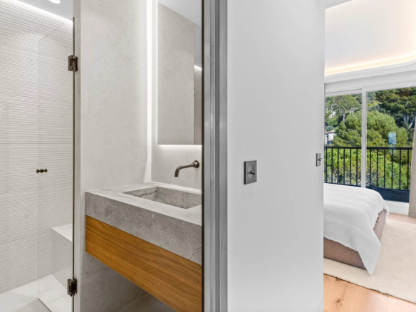 Contemporary bathroom with a glass shower enclosure and concrete countertop sink, adjacent to a bedroom with a balcony and a green outdoor view.
