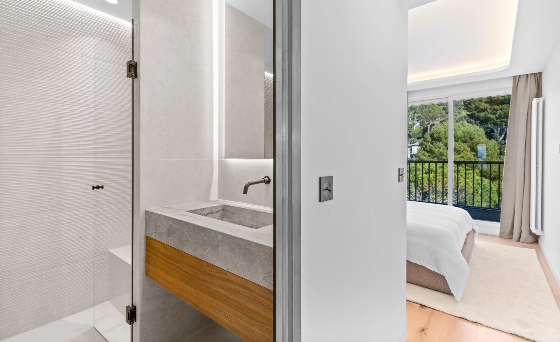 Contemporary bathroom with a glass shower enclosure and concrete countertop sink, adjacent to a bedroom with a balcony and a green outdoor view.