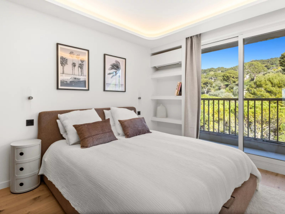 Bright bedroom with a large glass sliding door to a balcony and a view of green trees outside.
