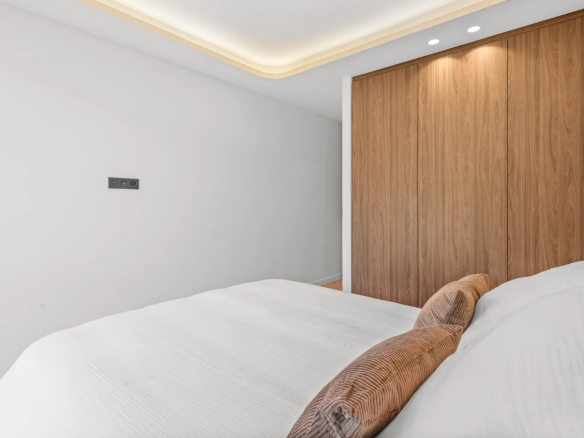Modern bedroom with white walls, a large wooden wardrobe, and a bed with white linens and brown cushions.