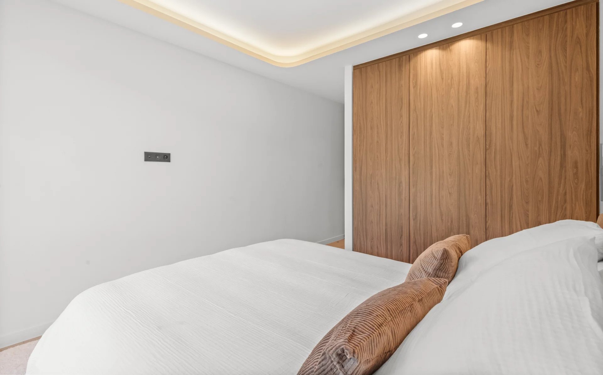 Modern bedroom with white walls, a large wooden wardrobe, and a bed with white linens and brown cushions.