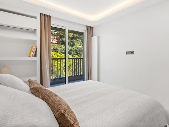 Minimalist white bedroom with a made bed, brown throw pillows, built-in shelves, and a balcony view of trees outside.