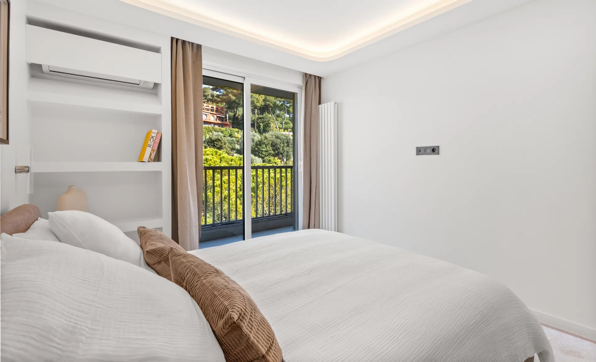 Minimalist white bedroom with a made bed, brown throw pillows, built-in shelves, and a balcony view of trees outside.