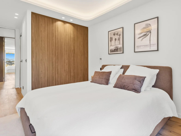 Bright modern bedroom with a large bed, white bedding, and a brown headboard beside a wooden built-in wardrobe; two framed palm prints on the wall.