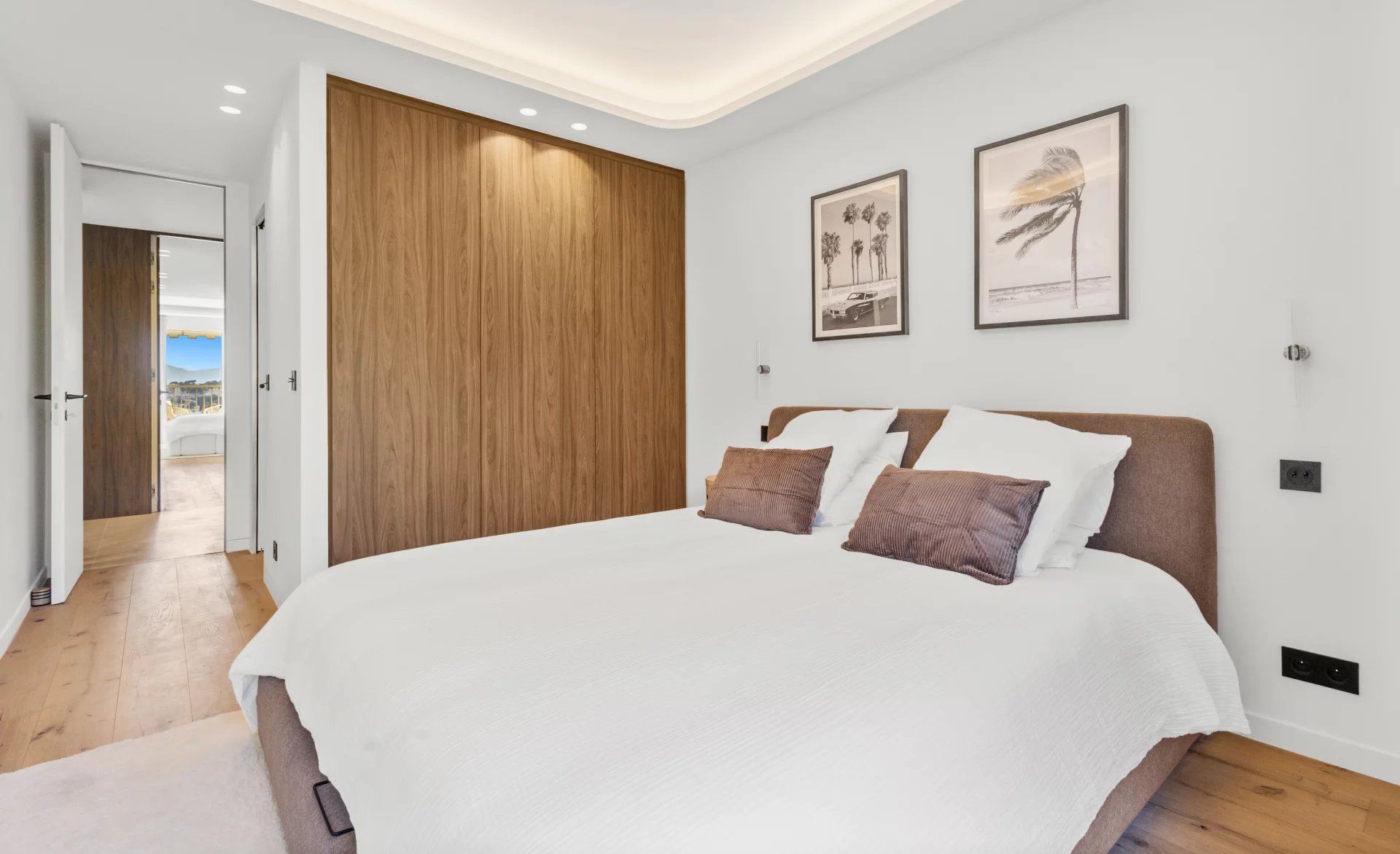 Bright modern bedroom with a large bed, white bedding, and a brown headboard beside a wooden built-in wardrobe; two framed palm prints on the wall.