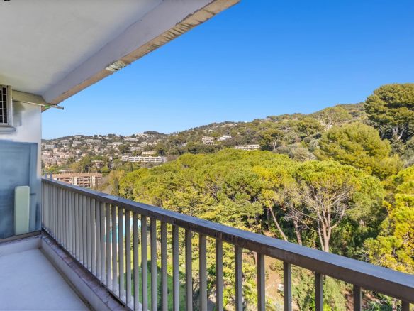 Balcony with metal railing overlooking a hillside neighborhood and dense green trees under a clear blue sky, with part of the building on the left.