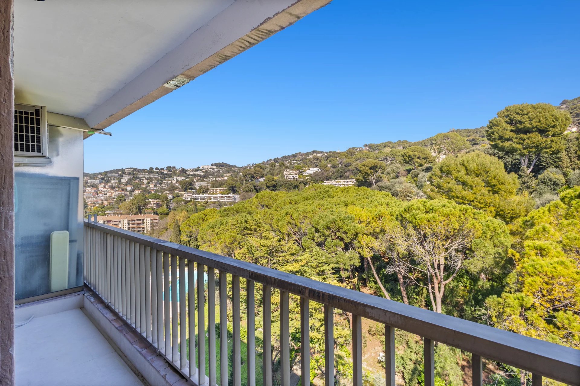 Balcony with metal railing overlooking a hillside neighborhood and dense green trees under a clear blue sky, with part of the building on the left.