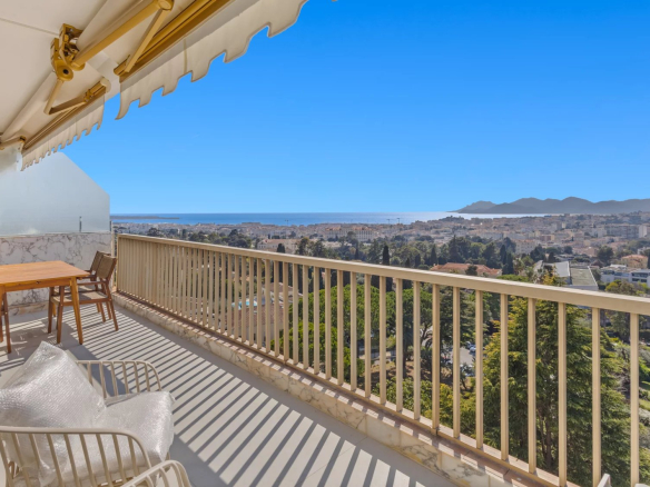 Balcony seating with a wooden table and chairs, overlooking a city, sea, and distant mountains under a clear blue sky.