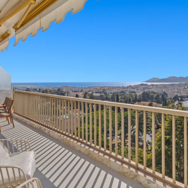 Balcony seating with a wooden table and chairs, overlooking a city, sea, and distant mountains under a clear blue sky.