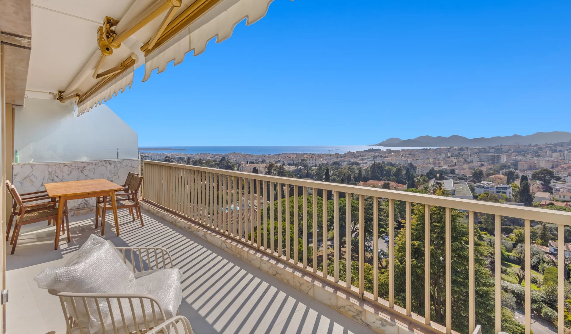Balcony seating with a wooden table and chairs, overlooking a city, sea, and distant mountains under a clear blue sky.