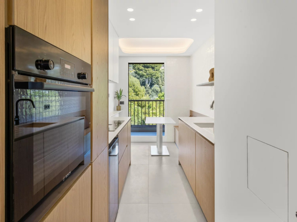 Modern galley kitchen with wood cabinets, a built-in black oven, and a small white table by a window overlooking greenery.