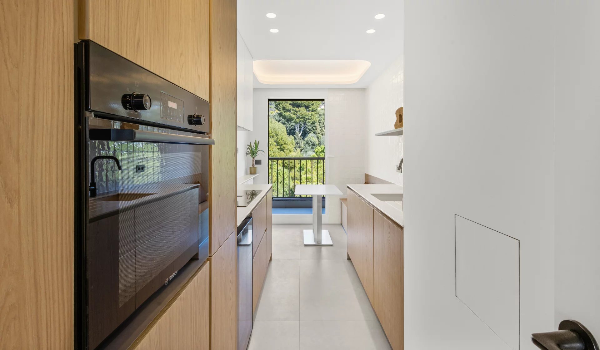 Modern galley kitchen with wood cabinets, a built-in black oven, and a small white table by a window overlooking greenery.